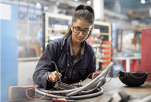 Eine Frau im Arbeitsoverall arbeitet mit einem Prüfgerät, dahinter eine Produktionshalle